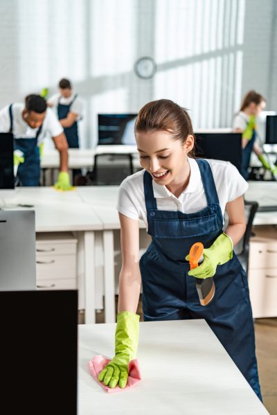 Female cleaner in overalls cleaning an office desk with a rag.